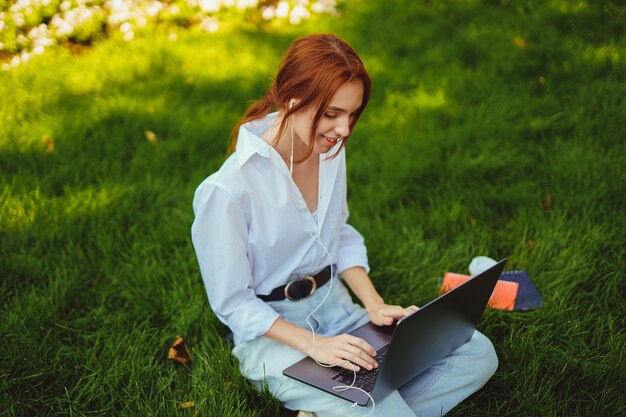 Bella giovane bella donna dai capelli rossi nel parco all'aperto utilizzando il computer portatile per studio o lavoro cablato...