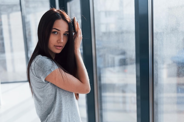 Bella giovane bruna in camicia grigia in piedi vicino alla finestra e in posa per una macchina fotografica.