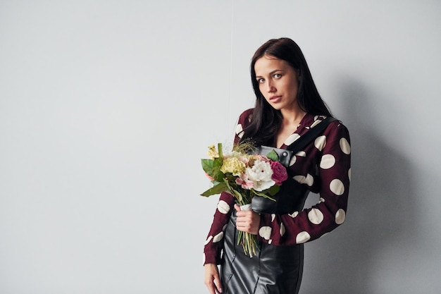 Bella giovane bruna in gonna nera in piedi al chiuso su sfondo bianco con bouquet in mano