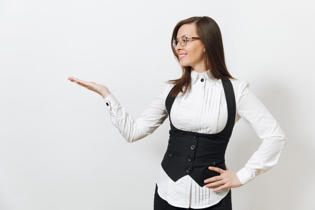 Bella giovane caucasica sorridente donna d'affari dai capelli castani in abito nero, camicia bianca e occhiali che puntano la mano da parte isolata sul muro bianco