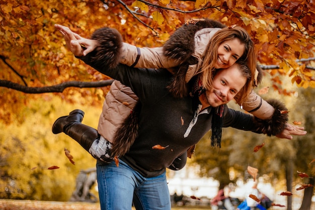 Bella giovane coppia godendo di un piggyback nel soleggiato parco nei colori autunnali. Guardando la fotocamera.