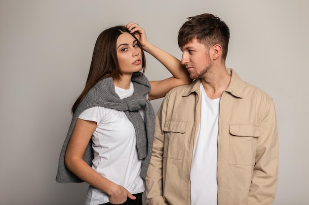 Bella giovane coppia in abiti alla moda con magliette bianche, maglioni vintage e una giacca beige in studio. Un uomo e una donna in capispalla casual posano su uno sfondo grigio