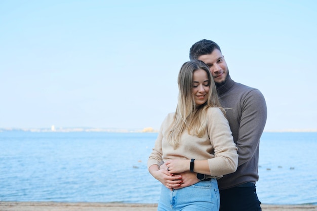 Bella giovane coppia in posa sulla spiaggia sullo sfondo di un grande lago Uomo che abbraccia la donna da dietro e guarda la macchina fotografica