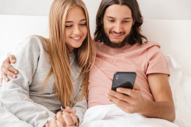 Bella giovane coppia seduta a letto a casa, guardando il cellulare