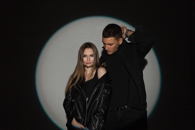 Bella giovane coppia uomo e donna in eleganti abiti neri casual in denim con una giacca di pelle e una camicia di jeans neri su uno sfondo scuro in un cerchio chiaro