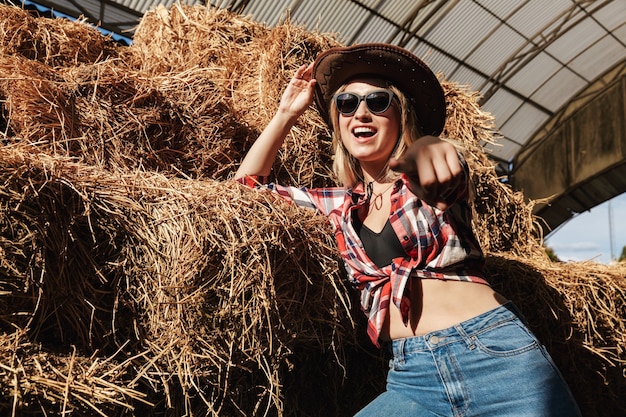 Bella giovane cowgirl bionda sorridente in piedi al fienile, appoggiata a un pagliaio