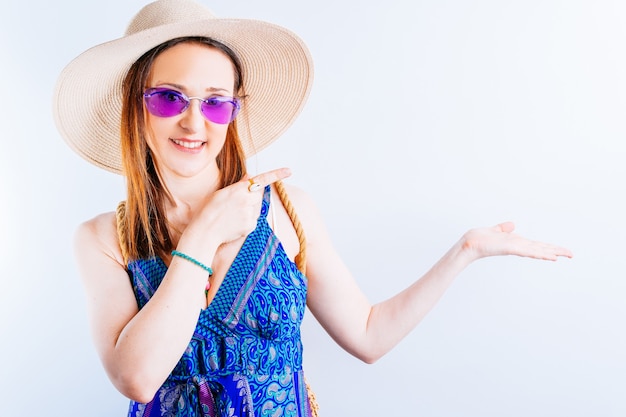 Bella giovane donna adulta che indossa cappello estivo, abito da spiaggia e occhiali viola in posa con la mano e puntando con il dito. commerciale o pubblicitario. concetto di estate