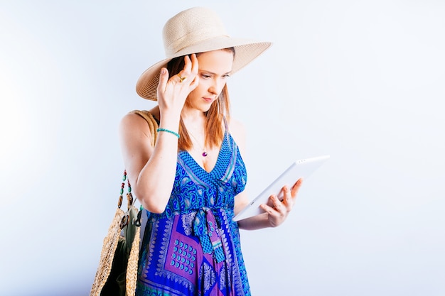 Bella giovane donna adulta in cappello da spiaggia e vestito guardando lo stato delle prenotazioni di viaggio sul tablet su sfondo bianco. concetto di organizzazione del viaggio. stato del volo