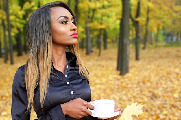 Bella giovane donna africana che beve caffè da una tazza bianca sullo sfondo di foglie di autunno nel parco