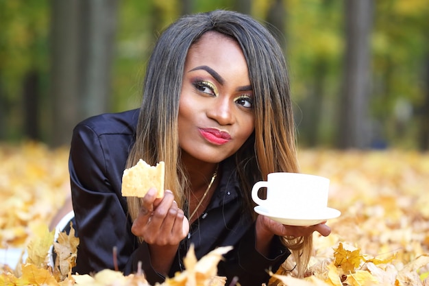 Bella giovane donna africana che beve caffè da una tazza bianca sullo spazio delle foglie di autunno nel parco