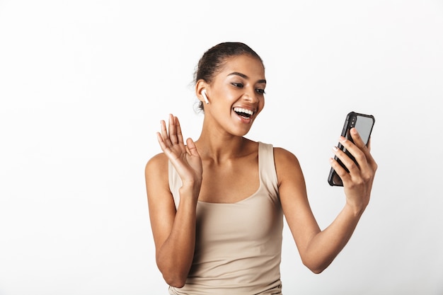 Bella giovane donna africana vestita casualmente in piedi isolato su bianco, indossando gli auricolari wireless, utilizzando il telefono cellulare