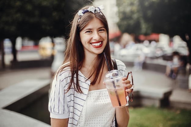 Bella giovane donna alla moda che tiene bevanda e si rilassa nella strada della città Felice ragazza hipster in abito retrò e fascia con bicchiere di plastica e cannuccia che beve limonata