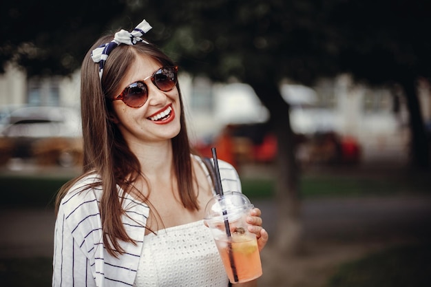 Bella giovane donna alla moda che tiene bevanda fredda e sorridente nella via della città Ragazza felice dei pantaloni a vita bassa con gli occhiali da sole in vestito retro che tiene la tazza e la paglia di plastica monouso