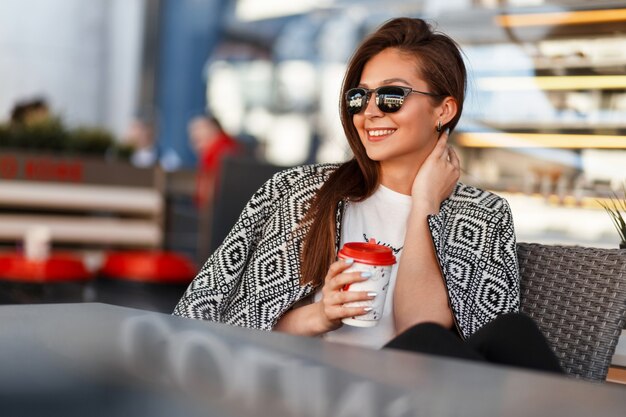 Bella giovane donna alla moda con caffè in occhiali da sole con vestiti alla moda alla moda