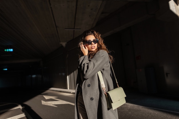 Bella giovane donna alla moda con capelli ricci e occhiali da sole alla moda in un cappotto lungo alla moda con una borsa vintage passeggiate in città. Stile e bellezza femminili urbani. Luce e ombra del sole