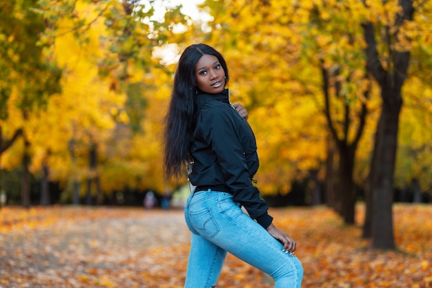 Bella giovane donna alla moda con la pelle nera in abiti alla moda con una giacca e jeans blu cammina in un parco autunnale con foglie autunnali di colore giallo brillante