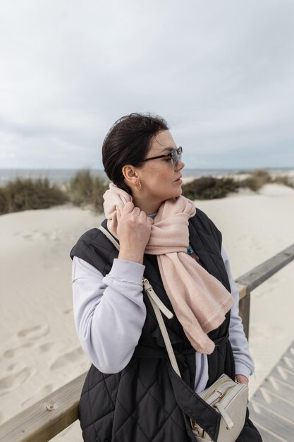 Bella giovane donna alla moda con occhiali da sole vintage che indossa capispalla primaverili alla moda con un gilet sciarpa rosa e una borsa pullover passeggiate sulla spiaggia in riva al mare