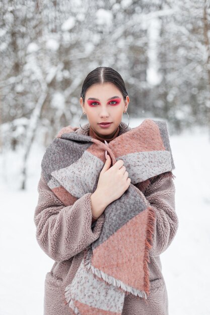 Bella giovane donna alla moda con trucco creativo colorato e ombretti rosa in abiti invernali alla moda con una sciarpa in posa nella foresta di neve