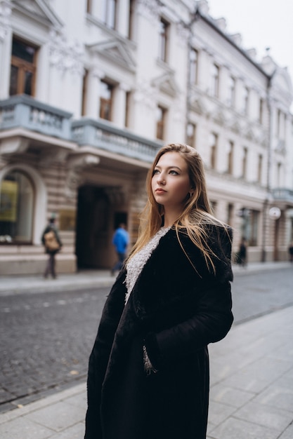 Bella giovane donna alla moda del blondie che posa in cappotto alla moda d'avanguardia all'aperto. Bellezza, moda, concetto di modellazione