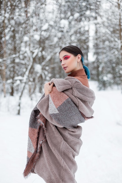 Bella giovane donna alla moda in abiti caldi invernali alla moda con una sciarpa e un piumino si trova nei boschi con la neve
