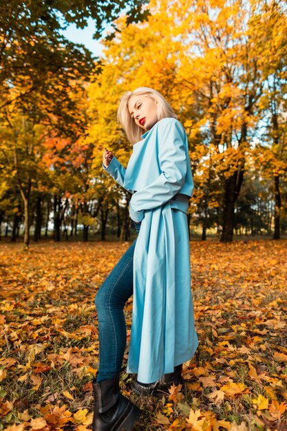 Bella giovane donna alla moda in cappotto blu alla moda con i jeans che posano in un parco di autunno con fogliame di colore giallo