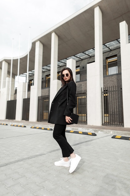 Bella giovane donna alla moda in elegante blazer nero con borsa vintage cammina vicino a un edificio moderno in città. La bellissima modella urbana della ragazza viaggia per strada. Abbigliamento casual giovanile alla moda per le donne.