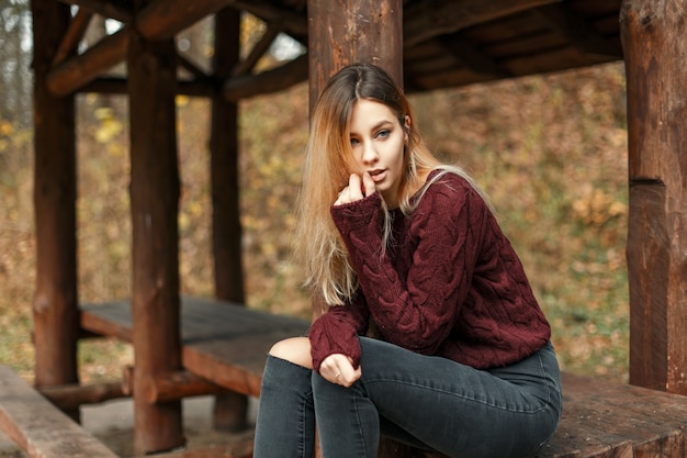 Bella giovane donna alla moda in jeans e maglione alla moda si siede in un pergolato in legno