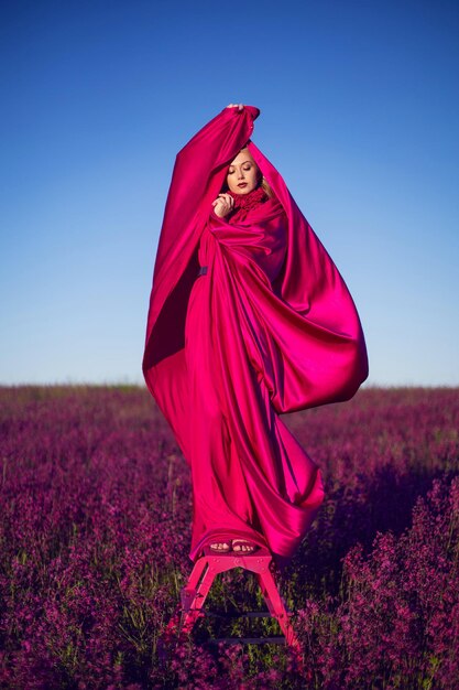 Bella giovane donna alla moda su una scala a pioli rosa in un vestito rosa in un campo con fiori di campo in estate al tramonto