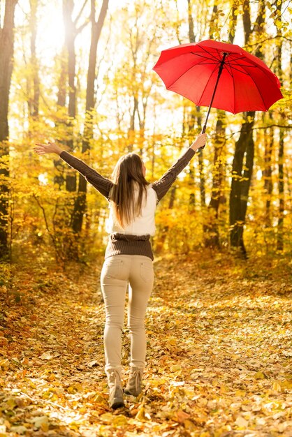 Bella giovane donna allegra con l'ombrello rosso divertendosi nella foresta soleggiata nei colori di autunno.