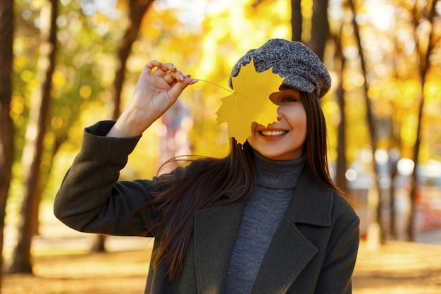 Bella giovane donna allegra con un sorriso in abiti vintage alla moda con un cappotto e un cappello con foglia di autunno giallo che cammina nel parco