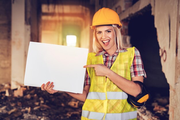 Bella giovane donna architetti edili nell'edificio danneggiato nel disastro. Indica una lavagna bianca vuota e sorride.