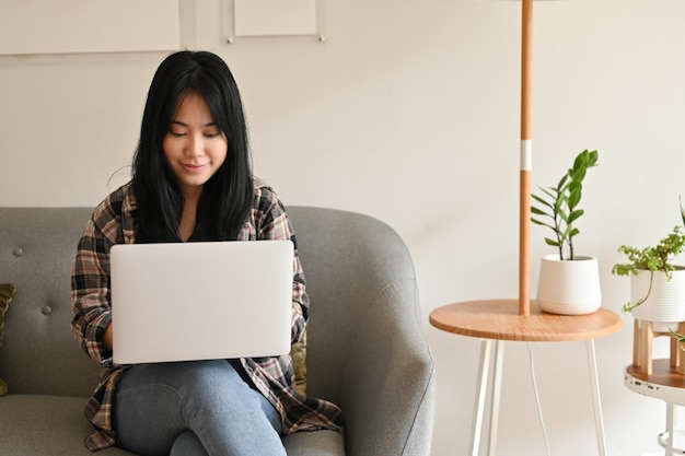 Bella giovane donna asiatica che lavora da casa utilizzando il computer portatile mentre si siede sul divano