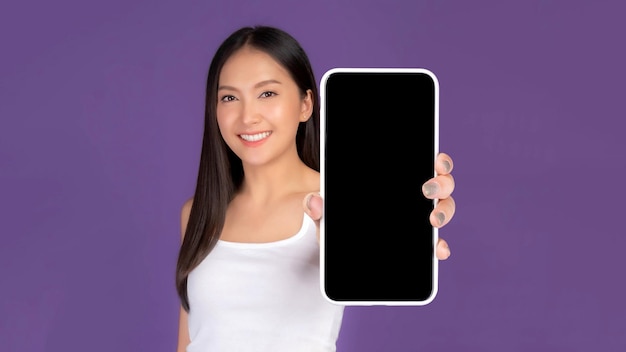 Bella giovane donna asiatica che mostra un grande smartphone con schermo bianco vuoto isolato su sfondo viola Visualizza Mock Up Image