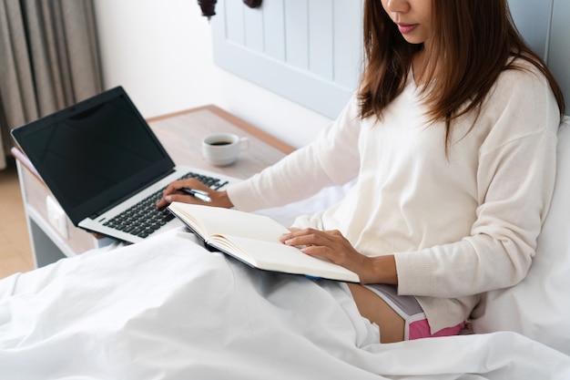 Bella giovane donna asiatica che si siede sul letto e che guarda il suo programma di tempo in taccuino mentre per mezzo del computer della compressa, lavorando sul letto, lavoro a casa, concetto occupato di stile di vita.