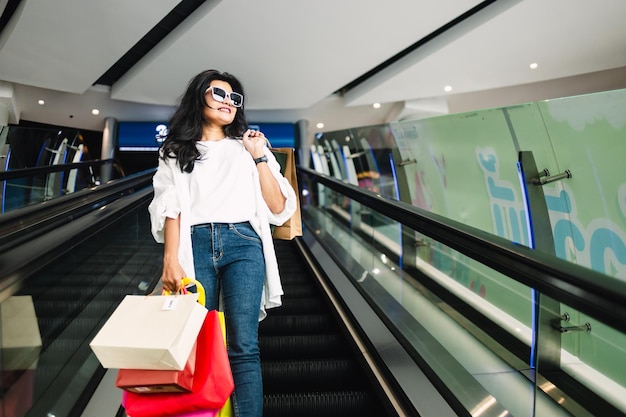 Bella giovane donna asiatica che tiene le borse della spesa e cammina sulla scala mobile nel centro commerciale
