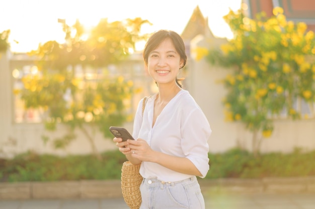 Bella giovane donna asiatica del ritratto con il telefono cellulare astuto intorno alla vista all'aperto della via in un giorno di estate