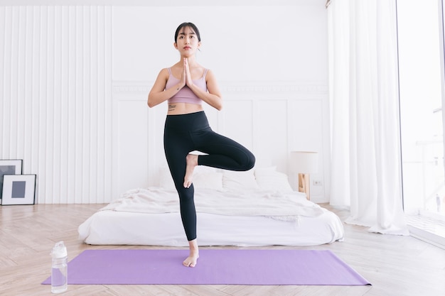 Bella giovane donna asiatica felice in piedi nella posizione di Vrksasana facendo esercizio di yoga sul tappetino viola con bottiglia d'acqua che si allena in camera da letto allenamento indoor Tree Pose a figura intera