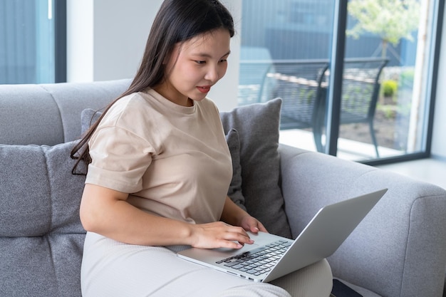 bella giovane donna asiatica in abiti casual e accoglienti utilizzando il computer portatile seduto sul divano in un appartamento moderno