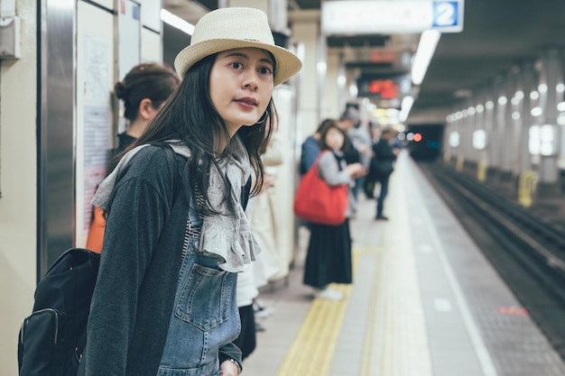 Bella giovane donna asiatica in attesa del treno sulla piattaforma della stazione della metropolitana. zaino in spalla femminile con cappello di paglia cercando ferrovia della metropolitana. le persone si spostano in viaggio concetto pubblico stile di vita.