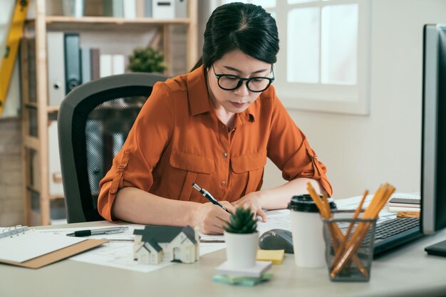 bella giovane donna asiatica ingegnere lavoratore che lavora a casa in ufficio su progetto. designer d'interni ragazza seduta al tavolo bianco che scrive idee creative su carte con una tazza di caffè sulla scrivania. signora architetto disegnare.