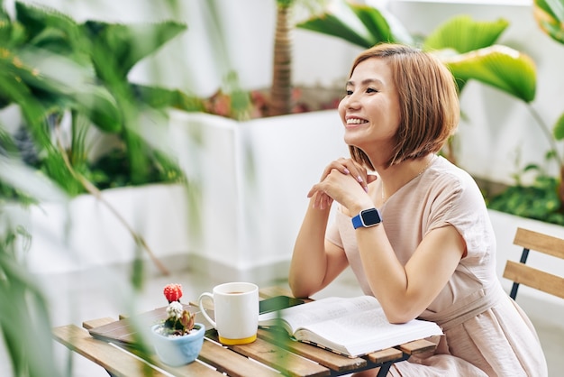 Bella giovane donna asiatica sorridente che si siede al tavolino del bar con il libro aperto, tablet PC e tazza di caffè