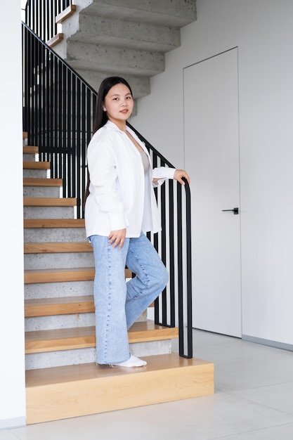 bella giovane donna asiatica sorridente con lunghi capelli scuri in camicia bianca sulle scale all'interno moderno