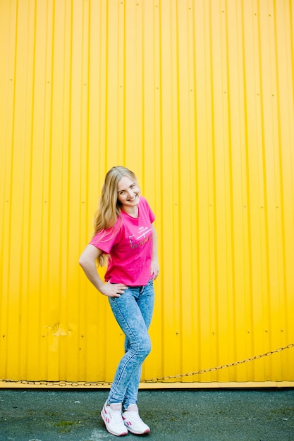 Bella giovane donna atletica con capelli biondi lunghi in una camicia rosa e blue jeans. In scarpe da ginnastica bianche. In posa e sorridendo alla parete del garage sulla parete gialla Posto per il testo. Le mani sulla cintura