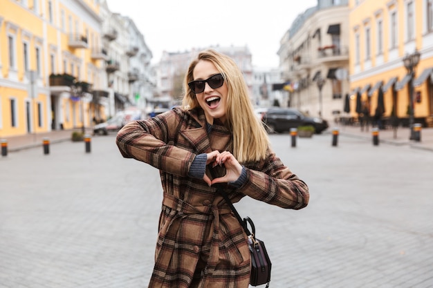 Bella giovane donna bionda alla moda che indossa un cappotto che cammina all'aperto, mostrando il gesto del cuore