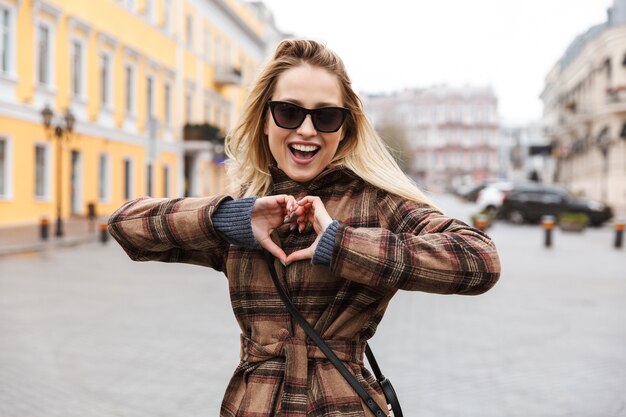 Bella giovane donna bionda alla moda che indossa un cappotto che cammina all'aperto, mostrando il gesto del cuore