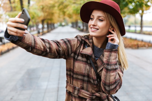 Bella giovane donna bionda alla moda che indossa un cappotto che cammina all'aperto, prendendo un selfie con il telefono cellulare