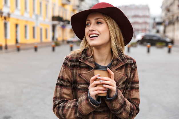 Bella giovane donna bionda alla moda che indossa un cappotto che cammina all'aperto, tenendo la tazza di caffè da asporto