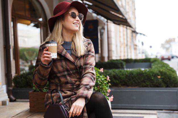 Bella giovane donna bionda alla moda che indossa un cappotto che si distende all'aperto, che tiene tazza da asporto