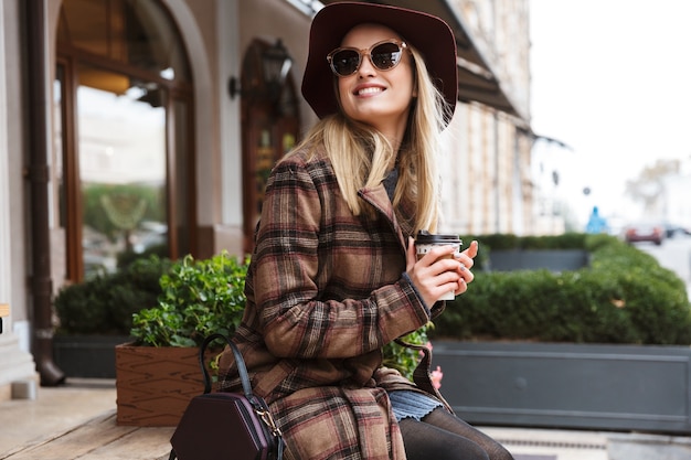 Bella giovane donna bionda alla moda che indossa un cappotto che si distende all'aperto, che tiene tazza da asporto