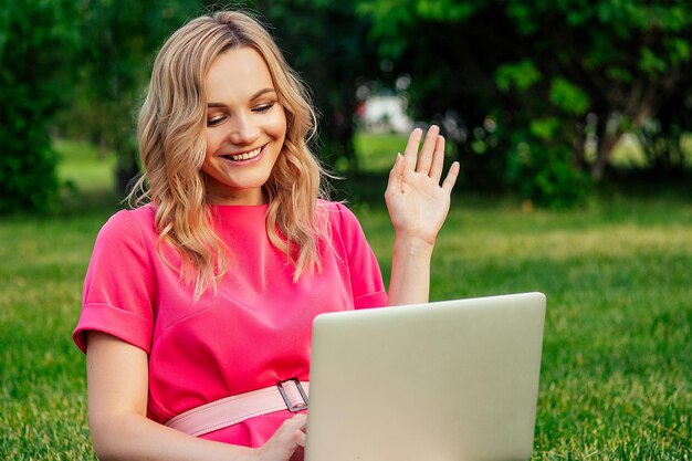 Bella giovane donna bionda allegra attiva in un vestito rosa che si siede sul prato di erba che lavora e che usa laptopl nel parco estivo